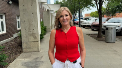A woman with a red shirt stands on a sidewalk and smiles at the camera.