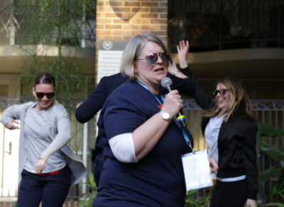 Marie speaks into a microphone, with people in the background.
